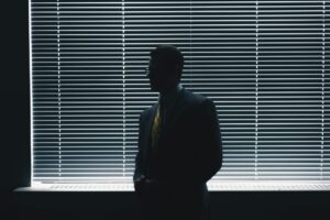 Un homme en costard-cravate est debout devant une fenêtre, la pièce est assombrie.