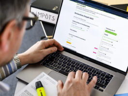 Changement de l’imposition sur les biens immobiliers : un cadeau pour les propriétaires les plus riches Gros plan sur les mains d’une personne tapant au clavier d’un MacBook Pro dont l’écran affiche le formulaire en ligne de déclaration d’impôt 2023 de l’État de Vaud. Sur le bureau, on distingue un classeur noir étiqueté “IMPÔTS”, un document imprimé partiellement visible et un surligneur jaune posés à côté du portable