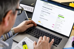Gros plan sur les mains d’une personne tapant au clavier d’un MacBook Pro dont l’écran affiche le formulaire en ligne de déclaration d’impôt 2023 de l’État de Vaud. Sur le bureau, on distingue un classeur noir étiqueté “IMPÔTS”, un document imprimé partiellement visible et un surligneur jaune posés à côté du portable