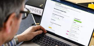 Gros plan sur les mains d’une personne tapant au clavier d’un MacBook Pro dont l’écran affiche le formulaire en ligne de déclaration d’impôt 2023 de l’État de Vaud. Sur le bureau, on distingue un classeur noir étiqueté “IMPÔTS”, un document imprimé partiellement visible et un surligneur jaune posés à côté du portable