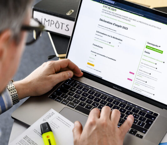 Changement de l’imposition sur les biens immobiliers : un cadeau pour les propriétaires les plus riches Gros plan sur les mains d’une personne tapant au clavier d’un MacBook Pro dont l’écran affiche le formulaire en ligne de déclaration d’impôt 2023 de l’État de Vaud. Sur le bureau, on distingue un classeur noir étiqueté “IMPÔTS”, un document imprimé partiellement visible et un surligneur jaune posés à côté du portable