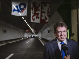 Albert Rösti : comment un conseiller fédéral impose son propre agenda Albert Rösti, en costume sombre, cravate bleue et lunettes, est interviewé au micro « KEYSTONE » à l’entrée d’un tunnel routier orné de banderoles (dont un panneau « A9 » et un drapeau à deux lions rouges) et d’une petite croix suisse suspendue au plafond.
