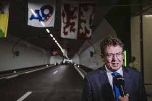 Albert Rösti, en costume sombre, cravate bleue et lunettes, est interviewé au micro « KEYSTONE » à l’entrée d’un tunnel routier orné de banderoles (dont un panneau « A9 » et un drapeau à deux lions rouges) et d’une petite croix suisse suspendue au plafond.