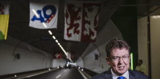 Albert Rösti, en costume sombre, cravate bleue et lunettes, est interviewé au micro « KEYSTONE » à l’entrée d’un tunnel routier orné de banderoles (dont un panneau « A9 » et un drapeau à deux lions rouges) et d’une petite croix suisse suspendue au plafond.
