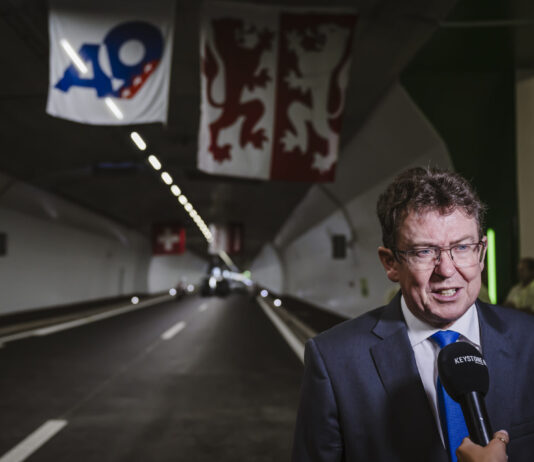 Albert Rösti : comment un conseiller fédéral impose son propre agenda Albert Rösti, en costume sombre, cravate bleue et lunettes, est interviewé au micro « KEYSTONE » à l’entrée d’un tunnel routier orné de banderoles (dont un panneau « A9 » et un drapeau à deux lions rouges) et d’une petite croix suisse suspendue au plafond.