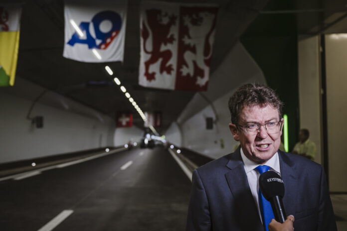 Albert Rösti, en costume sombre, cravate bleue et lunettes, est interviewé au micro « KEYSTONE » à l’entrée d’un tunnel routier orné de banderoles (dont un panneau « A9 » et un drapeau à deux lions rouges) et d’une petite croix suisse suspendue au plafond.