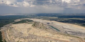 Vue aérienne d’une vaste carrière à ciel ouvert creusée en terrasses successives, avec un petit plan d’eau au fond. Les contours de l’excavation forment des courbes régulières, contrastant avec la ceinture de forêt et de champs qui l’entourent, le tout sous un ciel nuageux.