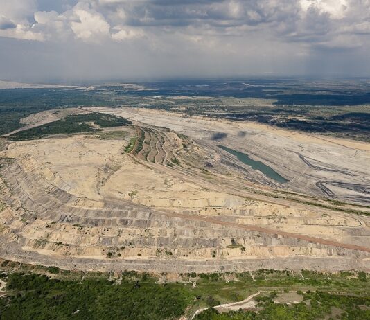 Glencore au défi de fermer la mine de Prodeco en Colombie Vue aérienne d’une vaste carrière à ciel ouvert creusée en terrasses successives, avec un petit plan d’eau au fond. Les contours de l’excavation forment des courbes régulières, contrastant avec la ceinture de forêt et de champs qui l’entourent, le tout sous un ciel nuageux.