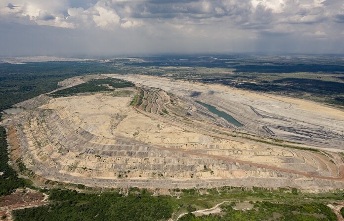 Vue aérienne d’une vaste carrière à ciel ouvert creusée en terrasses successives, avec un petit plan d’eau au fond. Les contours de l’excavation forment des courbes régulières, contrastant avec la ceinture de forêt et de champs qui l’entourent, le tout sous un ciel nuageux.