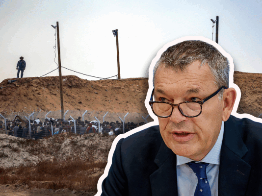 Philippe Lazzarini : « Les centres de la GHF sont des roulettes russes, un système cruel » Image composée en deux plans juxtaposés. À l’arrière, on distingue un terrain sombre et aride bordé d’un talus de terre surmonté de lampadaires et d’un soldat de la GHF en position de surveillance. Au pied de la butte, une clôture métallique coiffée de barbelés sépare un groupe dense de personnes du paysage extérieur. En surimpression à droite, découpé sur fond blanc, Philippe Lazzarini, un homme d’âge moyen en costume sombre, cravate et lunettes regarde vers sa gauche comme en pleine prise de parole, assis devant un pupitre ou un bureau.