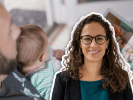 « Le congé parental doit offrir du temps supplémentaire sans compromettre la protection des mères » selon une experte Une femme souriante aux cheveux bouclés et portant des lunettes apparaît en surimpression sur la droite de l’image. À l’arrière-plan, une personne adulte tient un bébé sur ses genoux et lui lit un livre illustré dans une pièce lumineuse.