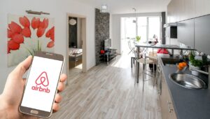 Une main tenant un smartphone affichant le logo d’Airbnb au premier plan, avec en arrière-plan un appartement moderne et lumineux comprenant une cuisine équipée, un salon avec télévision, une table à manger et une chambre visible par une porte ouverte. Une toile décorative représentant des fleurs rouges est accrochée au mur.