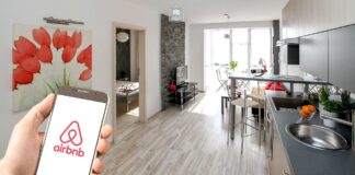 Une main tenant un smartphone affichant le logo d’Airbnb au premier plan, avec en arrière-plan un appartement moderne et lumineux comprenant une cuisine équipée, un salon avec télévision, une table à manger et une chambre visible par une porte ouverte. Une toile décorative représentant des fleurs rouges est accrochée au mur.