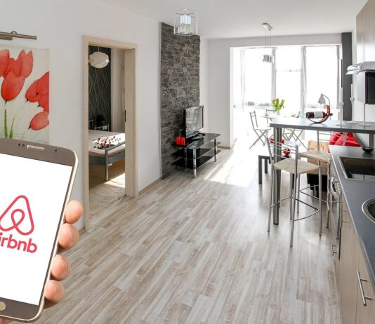 Nouvelles règles pour Airbnb : les sites touristiques européens agissent Une main tenant un smartphone affichant le logo d’Airbnb au premier plan, avec en arrière-plan un appartement moderne et lumineux comprenant une cuisine équipée, un salon avec télévision, une table à manger et une chambre visible par une porte ouverte. Une toile décorative représentant des fleurs rouges est accrochée au mur.