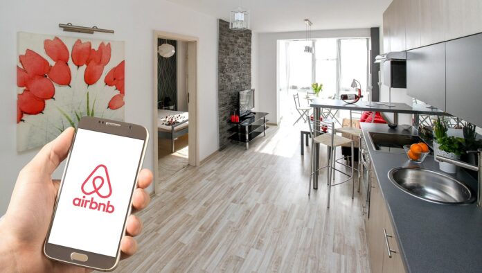 Une main tenant un smartphone affichant le logo d’Airbnb au premier plan, avec en arrière-plan un appartement moderne et lumineux comprenant une cuisine équipée, un salon avec télévision, une table à manger et une chambre visible par une porte ouverte. Une toile décorative représentant des fleurs rouges est accrochée au mur.