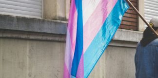 Un drapeau de la fierté transgenre aux bandes horizontales bleu clair, rose et blanc flotte sur une hampe en bois, tenu par une personne vue de dos en veste en jean, devant un mur de pierre grise usée.