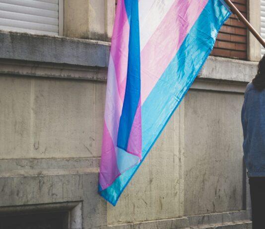 Attaque contre les personnes trans : l’UDC ignore le consensus scientifique Un drapeau de la fierté transgenre aux bandes horizontales bleu clair, rose et blanc flotte sur une hampe en bois, tenu par une personne vue de dos en veste en jean, devant un mur de pierre grise usée.