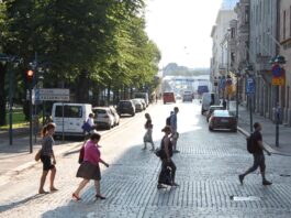 Helsinki : une année sans aucun mort sur la route grâce aux zones 30 km/h Plusieurs piéton-nes traversent à pied un large boulevard pavé baigné de lumière en fin d’après-midi à Helsinki, en Finlande. Au premier plan, cinq personnes marchent dans différentes directions. À gauche, derrière une rangée de voitures stationnées, de grands arbres forment un espace ombragé encadré par un panneau de circulation indiquant « Kluuvi » et « Kasarmitori ». À droite, des façades de bâtiments historiques et d’autres véhicules en stationnement bordent la rue. L’avenue s’ouvre vers un horizon lumineux où l’on distingue encore quelques automobilistes et passant-es.