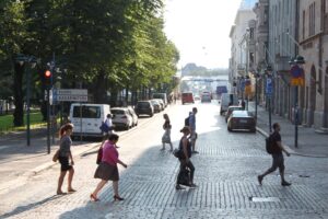 Plusieurs piéton-nes traversent à pied un large boulevard pavé baigné de lumière en fin d’après-midi à Helsinki, en Finlande. Au premier plan, cinq personnes marchent dans différentes directions. À gauche, derrière une rangée de voitures stationnées, de grands arbres forment un espace ombragé encadré par un panneau de circulation indiquant « Kluuvi » et « Kasarmitori ». À droite, des façades de bâtiments historiques et d’autres véhicules en stationnement bordent la rue. L’avenue s’ouvre vers un horizon lumineux où l’on distingue encore quelques automobilistes et passant-es.