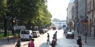 Plusieurs piéton-nes traversent à pied un large boulevard pavé baigné de lumière en fin d’après-midi à Helsinki, en Finlande. Au premier plan, cinq personnes marchent dans différentes directions. À gauche, derrière une rangée de voitures stationnées, de grands arbres forment un espace ombragé encadré par un panneau de circulation indiquant « Kluuvi » et « Kasarmitori ». À droite, des façades de bâtiments historiques et d’autres véhicules en stationnement bordent la rue. L’avenue s’ouvre vers un horizon lumineux où l’on distingue encore quelques automobilistes et passant-es.