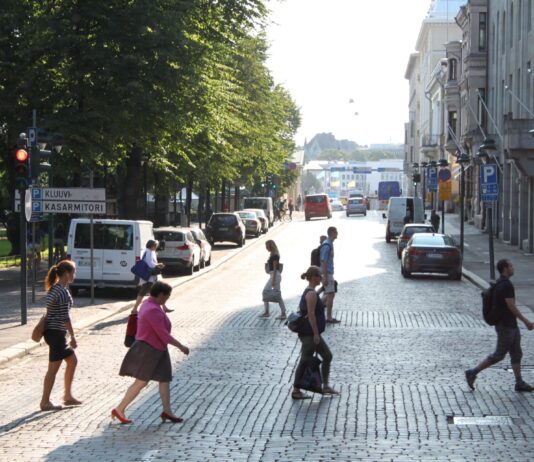 Helsinki : une année sans aucun mort sur la route grâce aux zones 30 km/h Plusieurs piéton-nes traversent à pied un large boulevard pavé baigné de lumière en fin d’après-midi à Helsinki, en Finlande. Au premier plan, cinq personnes marchent dans différentes directions. À gauche, derrière une rangée de voitures stationnées, de grands arbres forment un espace ombragé encadré par un panneau de circulation indiquant « Kluuvi » et « Kasarmitori ». À droite, des façades de bâtiments historiques et d’autres véhicules en stationnement bordent la rue. L’avenue s’ouvre vers un horizon lumineux où l’on distingue encore quelques automobilistes et passant-es.