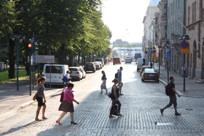 Plusieurs piéton-nes traversent à pied un large boulevard pavé baigné de lumière en fin d’après-midi à Helsinki, en Finlande. Au premier plan, cinq personnes marchent dans différentes directions. À gauche, derrière une rangée de voitures stationnées, de grands arbres forment un espace ombragé encadré par un panneau de circulation indiquant « Kluuvi » et « Kasarmitori ». À droite, des façades de bâtiments historiques et d’autres véhicules en stationnement bordent la rue. L’avenue s’ouvre vers un horizon lumineux où l’on distingue encore quelques automobilistes et passant-es.