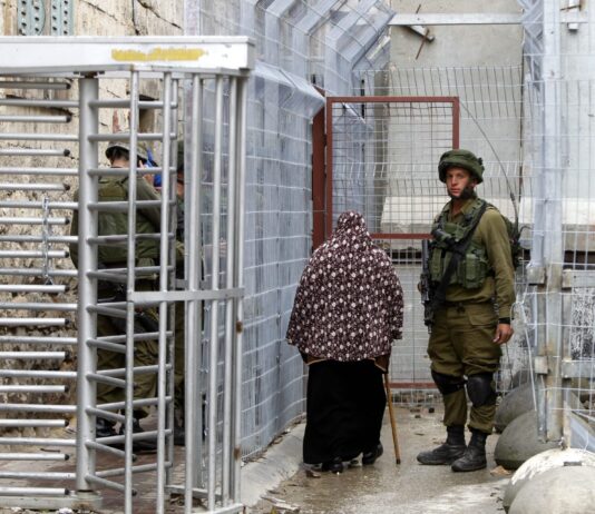 Pression sur Israël : pourquoi la solution à deux États est importante Une femme voilée de dos, portant un long vêtement noir et un châle à motifs floraux marron, avance à l’aide d’une canne dans un passage étroit bordé de grillages métalliques. À gauche, un tourniquet en acier et, derrière, un soldat en uniforme kaki et casque, partiellement masqué. À droite, un autre soldat en tenue de combat kaki, casque sur la tête et fusil en bandoulière, se tient face à l’objectif. Le sol est mouillé et des blocs de béton renforcent la barrière de chaque côté.