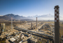 La Suisse continue de chuter dans le classement climatique Vue aérienne d’un vaste complexe industriel posé dans une plaine bordée de montagnes enneigées sous un ciel bleu. Au premier plan, de nombreux bâtiments, réseaux de tuyauteries et plateformes métalliques s’étendent horizontalement. Trois grandes cheminées – dont l’une ornée d’un damier blanc et rouge – dominent l’ensemble. À l’arrière-plan, on aperçoit des champs ouverts et, à l’horizon, une chaîne de sommets alpins encore partiellement enneigés.