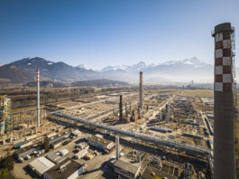 La Suisse continue de chuter dans le classement climatique Vue aérienne d’un vaste complexe industriel posé dans une plaine bordée de montagnes enneigées sous un ciel bleu. Au premier plan, de nombreux bâtiments, réseaux de tuyauteries et plateformes métalliques s’étendent horizontalement. Trois grandes cheminées – dont l’une ornée d’un damier blanc et rouge – dominent l’ensemble. À l’arrière-plan, on aperçoit des champs ouverts et, à l’horizon, une chaîne de sommets alpins encore partiellement enneigés.