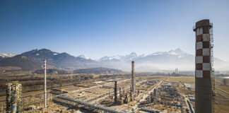 Vue aérienne d’un vaste complexe industriel posé dans une plaine bordée de montagnes enneigées sous un ciel bleu. Au premier plan, de nombreux bâtiments, réseaux de tuyauteries et plateformes métalliques s’étendent horizontalement. Trois grandes cheminées – dont l’une ornée d’un damier blanc et rouge – dominent l’ensemble. À l’arrière-plan, on aperçoit des champs ouverts et, à l’horizon, une chaîne de sommets alpins encore partiellement enneigés.