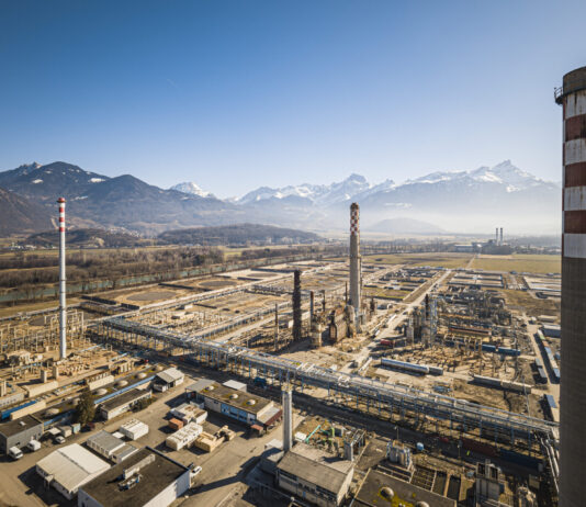 La Suisse continue de chuter dans le classement climatique Vue aérienne d’un vaste complexe industriel posé dans une plaine bordée de montagnes enneigées sous un ciel bleu. Au premier plan, de nombreux bâtiments, réseaux de tuyauteries et plateformes métalliques s’étendent horizontalement. Trois grandes cheminées – dont l’une ornée d’un damier blanc et rouge – dominent l’ensemble. À l’arrière-plan, on aperçoit des champs ouverts et, à l’horizon, une chaîne de sommets alpins encore partiellement enneigés.