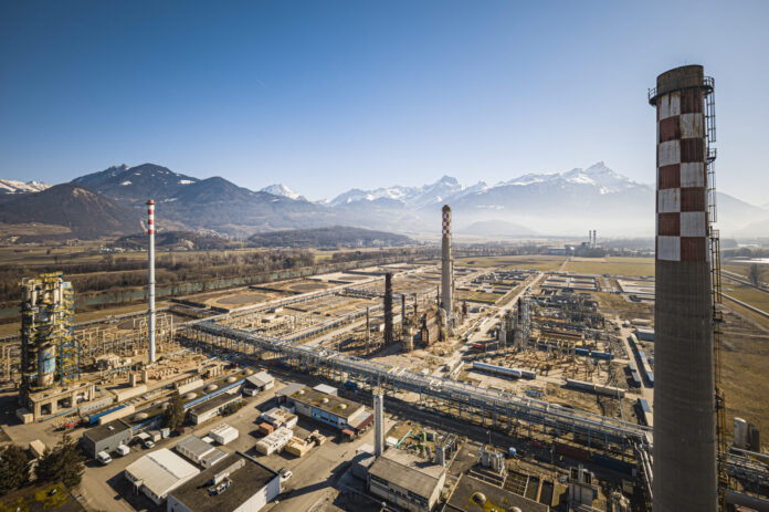 Vue aérienne d’un vaste complexe industriel posé dans une plaine bordée de montagnes enneigées sous un ciel bleu. Au premier plan, de nombreux bâtiments, réseaux de tuyauteries et plateformes métalliques s’étendent horizontalement. Trois grandes cheminées – dont l’une ornée d’un damier blanc et rouge – dominent l’ensemble. À l’arrière-plan, on aperçoit des champs ouverts et, à l’horizon, une chaîne de sommets alpins encore partiellement enneigés.