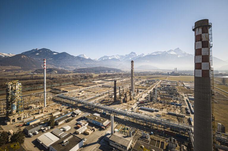 Vue aérienne d’un vaste complexe industriel posé dans une plaine bordée de montagnes enneigées sous un ciel bleu. Au premier plan, de nombreux bâtiments, réseaux de tuyauteries et plateformes métalliques s’étendent horizontalement. Trois grandes cheminées – dont l’une ornée d’un damier blanc et rouge – dominent l’ensemble. À l’arrière-plan, on aperçoit des champs ouverts et, à l’horizon, une chaîne de sommets alpins encore partiellement enneigés.