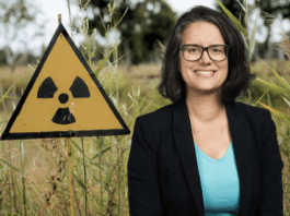 Oriane Sarrasin : « Le nucléaire, entre la peur et la foi technosolutionniste » Une femme portant des lunettes, un blazer noir et un T-shirt bleu clair sourit devant un panneau triangulaire jaune arborant le symbole radioactif, le tout planté dans des hautes herbes en milieu naturel.