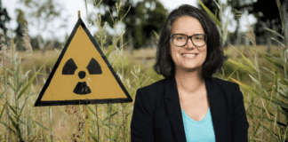 Une femme portant des lunettes, un blazer noir et un T-shirt bleu clair sourit devant un panneau triangulaire jaune arborant le symbole radioactif, le tout planté dans des hautes herbes en milieu naturel.