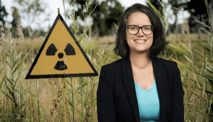 Une femme portant des lunettes, un blazer noir et un T-shirt bleu clair sourit devant un panneau triangulaire jaune arborant le symbole radioactif, le tout planté dans des hautes herbes en milieu naturel.