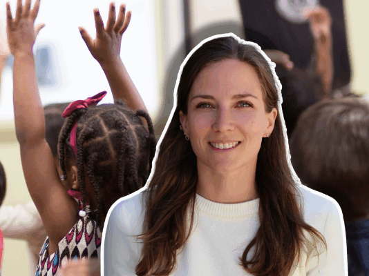 « La crèche : première école de la diversité » — Journal d’une éducatrice #4 Gros plan sur l’arrière d’une rangée d’enfants d’âges préscolaires, assis en classe et levant la main pour répondre à l’enseignant. L’un des enfants, une fillette aux cheveux tressés ornés de perles, porte une robe à motifs colorés. Le portrait d’une femme souriante aux cheveux bruns est superposé.