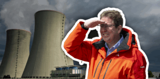 Albert Rösti, un homme en veste orange et lunettes se tient devant deux grandes tours de refroidissement d’une centrale électrique, levant la main au front pour regarder au loin sous un ciel gris et menaçant.