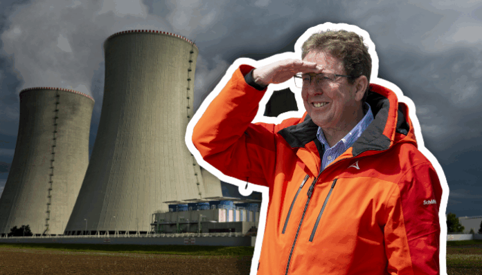 Albert Rösti, un homme en veste orange et lunettes se tient devant deux grandes tours de refroidissement d’une centrale électrique, levant la main au front pour regarder au loin sous un ciel gris et menaçant.