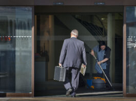 Écarts salariaux : les revenus des CEO augmentent de 18 % Photo couleur d’une entrée vitrée d’une agence UBS. Un homme d’affaires en costume, vu de dos, franchit le seuil en tenant une mallette noire dans une main et un journal dans l’autre. À droite, une employée de nettoyage en uniforme passe l’aspirateur sur le paillasson. Le logo UBS apparaît en rouge sur la porte en verre. Les écarts salariaux entre les CEO et les salarié-es continuent de se creuser.
