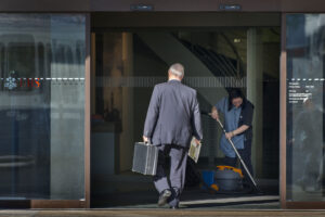 Photo couleur d’une entrée vitrée d’une agence UBS. Un homme d’affaires en costume, vu de dos, franchit le seuil en tenant une mallette noire dans une main et un journal dans l’autre. À droite, une employée de nettoyage en uniforme passe l’aspirateur sur le paillasson. Le logo UBS apparaît en rouge sur la porte en verre. Les écarts salariaux entre les CEO et les salarié-es continuent de se creuser.