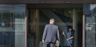 Photo couleur d’une entrée vitrée d’une agence UBS. Un homme d’affaires en costume, vu de dos, franchit le seuil en tenant une mallette noire dans une main et un journal dans l’autre. À droite, une employée de nettoyage en uniforme passe l’aspirateur sur le paillasson. Le logo UBS apparaît en rouge sur la porte en verre. Les écarts salariaux entre les CEO et les salarié-es continuent de se creuser.
