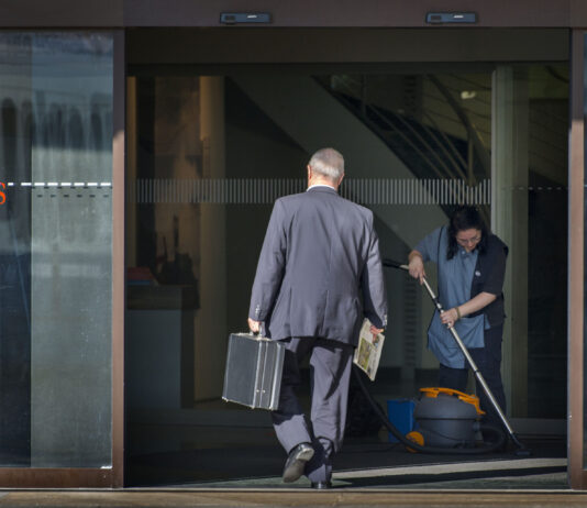Écarts salariaux : les revenus des CEO augmentent de 18 % Photo couleur d’une entrée vitrée d’une agence UBS. Un homme d’affaires en costume, vu de dos, franchit le seuil en tenant une mallette noire dans une main et un journal dans l’autre. À droite, une employée de nettoyage en uniforme passe l’aspirateur sur le paillasson. Le logo UBS apparaît en rouge sur la porte en verre. Les écarts salariaux entre les CEO et les salarié-es continuent de se creuser.