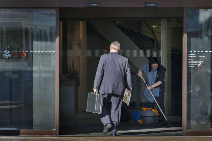 Photo couleur d’une entrée vitrée d’une agence UBS. Un homme d’affaires en costume, vu de dos, franchit le seuil en tenant une mallette noire dans une main et un journal dans l’autre. À droite, une employée de nettoyage en uniforme passe l’aspirateur sur le paillasson. Le logo UBS apparaît en rouge sur la porte en verre. Les écarts salariaux entre les CEO et les salarié-es continuent de se creuser.