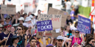 Vue d’une manifestation en plein air où un groupe divers de personnes marche dans la rue. Au centre, une jeune femme en haut violet lève une pancarte « My body, my choice ». Plusieurs manifestant·e·s autour portent des vêtements et accessoires violets et tiennent d’autres slogans (dont, en arrière-plan, « Things that cause rape : »). L’ambiance est estivale, la foule avance unie pour défendre les droits à l’avortement et l’autodétermination.