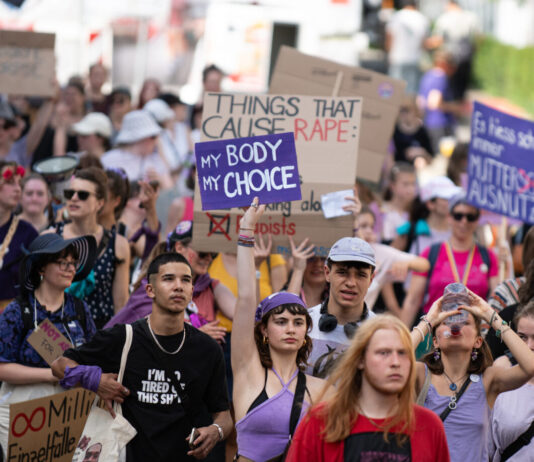 Bonnes nouvelles en septembre, de l’avortement pris en charge à l’atmosphère terrestre Vue d’une manifestation en plein air où un groupe divers de personnes marche dans la rue. Au centre, une jeune femme en haut violet lève une pancarte « My body, my choice ». Plusieurs manifestant·e·s autour portent des vêtements et accessoires violets et tiennent d’autres slogans (dont, en arrière-plan, « Things that cause rape : »). L’ambiance est estivale, la foule avance unie pour défendre les droits à l’avortement et l’autodétermination.