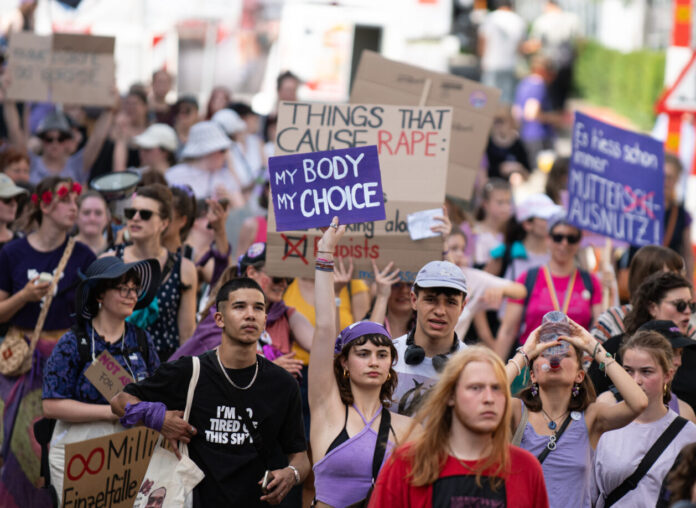 Vue d’une manifestation en plein air où un groupe divers de personnes marche dans la rue. Au centre, une jeune femme en haut violet lève une pancarte « My body, my choice ». Plusieurs manifestant·e·s autour portent des vêtements et accessoires violets et tiennent d’autres slogans (dont, en arrière-plan, « Things that cause rape : »). L’ambiance est estivale, la foule avance unie pour défendre les droits à l’avortement et l’autodétermination.