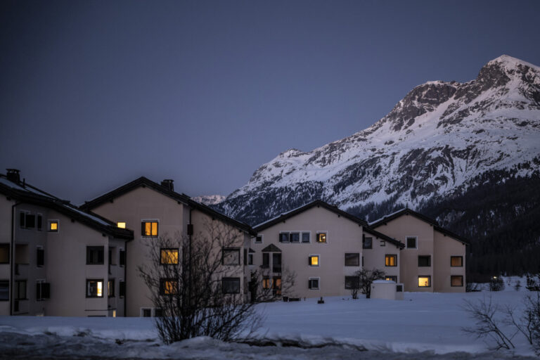 Vue de nuit tombante d’un petit ensemble de maisons beiges aux toits foncés à Sils, aux Grisons, dont plusieurs fenêtres sont baignées d’une lumière chaude. Elles se dressent au-dessus d’un champ enneigé, devant le flanc d’une haute montagne également couverte de neige, sous un ciel bleu sombre de crépuscule. Le canton des Grisons met en garde contre des hausses d'impôts entraînée par la suppression de la valeur locative.