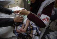 Gaza : ces organisations humanitaires sont toujours actives Bébé nouveau-né couché sur une table d’examen à Gaza, enveloppé dans une couverture à carreaux marron et bleu. Une soignante portant un gilet blanc et bordeaux marqué du logo Médecins Sans Frontières tient délicatement l’enfant par le torse et les épaules pour ajuster ses vêtements. On voit également les mains d’un autre adulte qui soutiennent doucement la tête du bébé. À l’arrière-plan, un évier fixé au mur et, à droite, une trousse médicale posée sur un meuble.