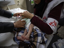 Gaza : ces organisations humanitaires sont toujours actives Bébé nouveau-né couché sur une table d’examen à Gaza, enveloppé dans une couverture à carreaux marron et bleu. Une soignante portant un gilet blanc et bordeaux marqué du logo Médecins Sans Frontières tient délicatement l’enfant par le torse et les épaules pour ajuster ses vêtements. On voit également les mains d’un autre adulte qui soutiennent doucement la tête du bébé. À l’arrière-plan, un évier fixé au mur et, à droite, une trousse médicale posée sur un meuble.